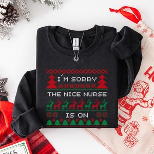 May include: Black crewneck sweatshirt with a festive Christmas design. The text reads "I'M SORRY THE NICE NURSE IS ON" in red and green. A silver necklace with a sun pendant is also visible.
