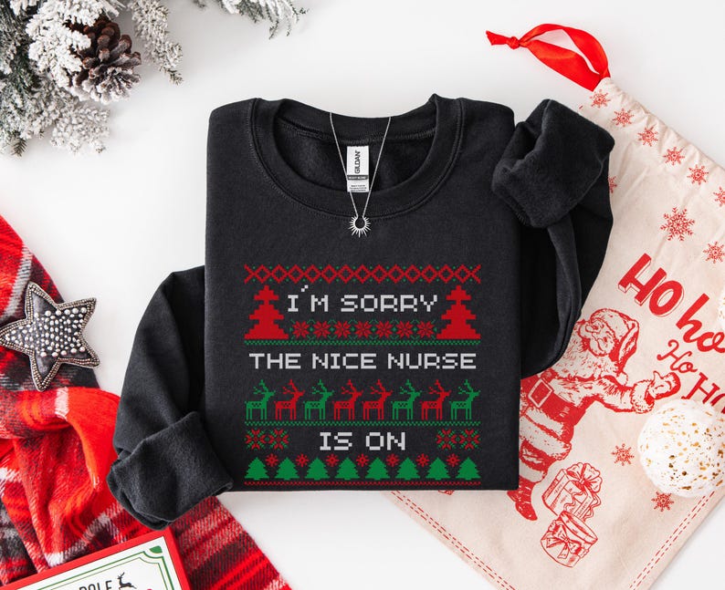 May include: Black crewneck sweatshirt with a festive Christmas design. The text reads "I'M SORRY THE NICE NURSE IS ON" in red and green. A silver necklace with a sun pendant is also visible.