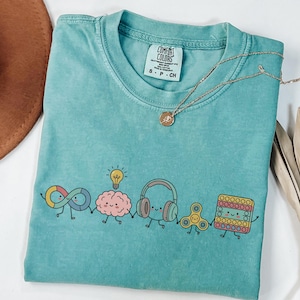 May include: A teal short-sleeved t-shirt featuring a graphic of five cartoon characters holding hands: an infinity symbol, a brain with a lightbulb, headphones, a fidget spinner, and a square pop-it toy. The shirt has a Comfort Colors label.