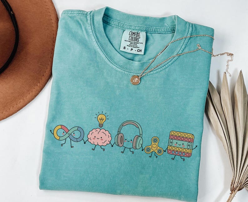 May include: A teal short-sleeved t-shirt featuring a graphic of five cartoon characters holding hands: an infinity symbol, a brain with a lightbulb, headphones, a fidget spinner, and a square pop-it toy. The shirt has a Comfort Colors label.