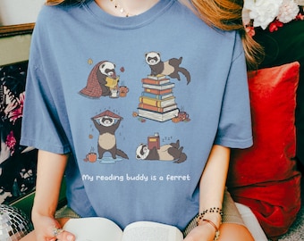 May include: A light blue t-shirt featuring a graphic of ferrets in various reading poses. The ferrets are depicted with books, a blanket, and a rain hat. The text "My reading buddy is a ferret" is printed below the graphic.
