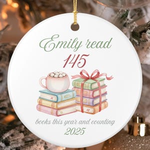 Puede incluir: Adorno navideño de cerámica blanca con una cuerda dorada. El adorno presenta el texto "Emily read 145" en escritura verde y roja. Debajo hay ilustraciones de libros y una taza de chocolate caliente. También se incluye el texto "books this year and counting 2025".