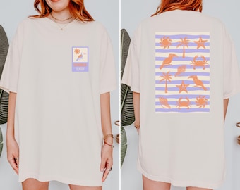 May include: Cream-colored t-shirt with a front and back design. The front features a square graphic with a bird and the text "Summer Season." The back has a rectangular design with beach-themed illustrations, including toucans, starfish, and crabs.