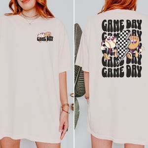 Op de afbeelding: Een oversized crèmekleurig t-shirt met een grafische print van twee cartoon-honkballen en een geruit patroon. De tekst "GAME DAY" wordt herhaald in een retro-lettertype op de achterkant van het shirt.