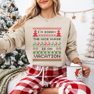 May include: Beige sweatshirt with a festive design. The text reads "I'M SORRY THE NICE NURSE IS ON VACATION" in a red and green pixelated pattern. The design includes reindeer and Christmas trees. The person is wearing red and white plaid pajama pants.