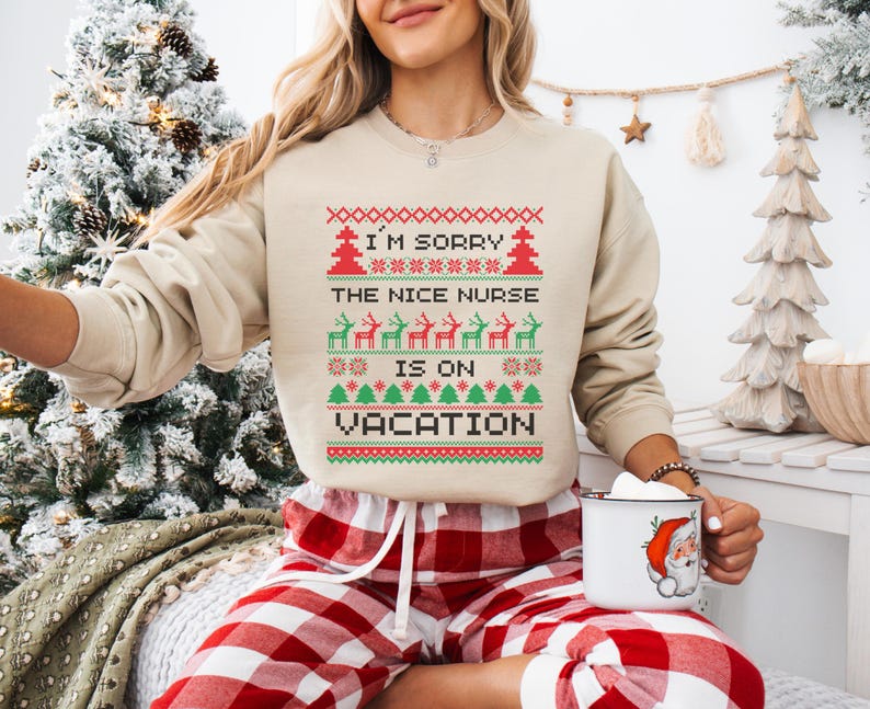 May include: Beige sweatshirt with a festive design. The text reads "I'M SORRY THE NICE NURSE IS ON VACATION" in a red and green pixelated pattern. The design includes reindeer and Christmas trees. The person is wearing red and white plaid pajama pants.