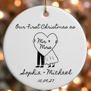 Puede incluir: Un adorno redondo de cerámica blanca con un dibujo lineal negro de una pareja sosteniendo un corazón que dice Mr & Mrs. El texto dice Our First Christmas as, Sophia + Michael y 10.09.27. Incluye un cordón dorado para colgar.