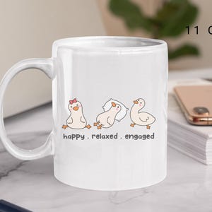 Könnte beinhalten: Weiße Keramiktasse mit einem skurrilen Design mit drei Cartoon-Enten in verschiedenen Posen, begleitet von den Worten "happy. relaxed. engaged." Die Tasse hat ein Fassungsvermögen von 325 ml.