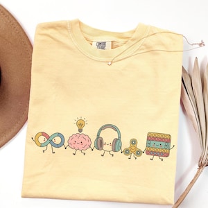 Comfort Colors Autism Doodle Characters Shirt featuring colorful, hand-drawn inclusive characters. Neatly styled next to classroom supplies, making it an ideal inclusion shirt, autism teacher shirt, and back to school shirt.