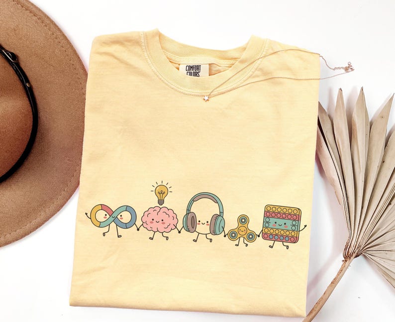 Comfort Colors Autism Doodle Characters Shirt featuring colorful, hand-drawn inclusive characters. Neatly styled next to classroom supplies, making it an ideal inclusion shirt, autism teacher shirt, and back to school shirt.