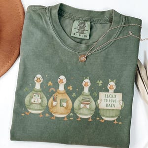 Puede incluir: Camiseta verde salvia con un diseño de cuatro gansos con suéteres, sosteniendo libros y lápices, y un letrero que dice "Lucky to Love Data". Un collar dorado y un sombrero marrón también están en la imagen.
