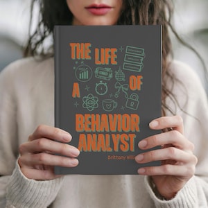 以下が含まれることがあります： オレンジと緑の文字で「THE LIFE OF A BEHAVIOR ANALYST」というタイトルが書かれた濃い灰色のノート。表紙には様々なアイコンと著者の名前が描かれています。ノートは人が持っています。