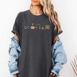 Comfort Colors Autism Doodle Characters Shirt featuring colorful, hand-drawn inclusive characters. Neatly styled next to classroom supplies, making it an ideal inclusion shirt, autism teacher shirt, and back to school shirt.