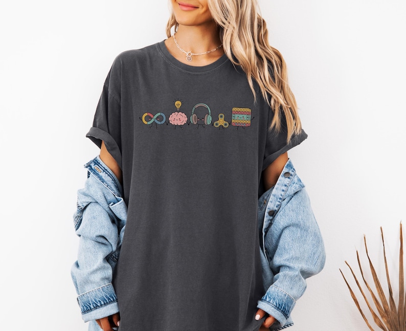 Comfort Colors Autism Doodle Characters Shirt featuring colorful, hand-drawn inclusive characters. Neatly styled next to classroom supplies, making it an ideal inclusion shirt, autism teacher shirt, and back to school shirt.