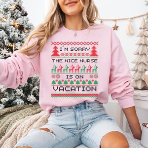 May include: A light pink sweatshirt with a festive design. The text "I'M SORRY THE NICE NURSE IS ON VACATION" is displayed in a pixelated style, along with Christmas trees and reindeer. The sweatshirt is a long-sleeved crew neck.