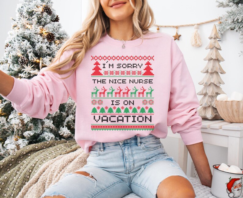 May include: A light pink sweatshirt with a festive design. The text "I'M SORRY THE NICE NURSE IS ON VACATION" is displayed in a pixelated style, along with Christmas trees and reindeer. The sweatshirt is a long-sleeved crew neck.