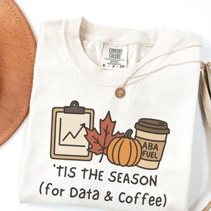 May include: Off-white t-shirt featuring a seasonal graphic. The design includes a clipboard with a graph, a maple leaf, a pumpkin, and a coffee cup with "ABA FUEL." The text reads "'TIS THE SEASON (for Data & Coffee)." A gold necklace is draped across the shirt.