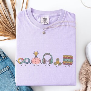 Comfort Colors Autism Doodle Characters Shirt featuring colorful, hand-drawn inclusive characters. Neatly styled next to classroom supplies, making it an ideal inclusion shirt, autism teacher shirt, and back to school shirt.