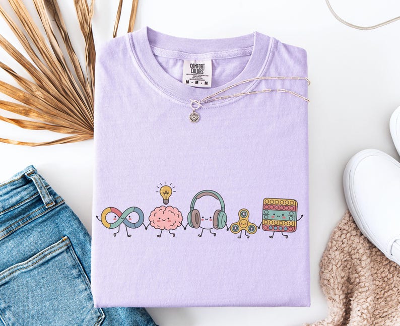 Comfort Colors Autism Doodle Characters Shirt featuring colorful, hand-drawn inclusive characters. Neatly styled next to classroom supplies, making it an ideal inclusion shirt, autism teacher shirt, and back to school shirt.