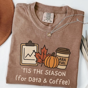 May include: Brown Comfort Colors t-shirt with a fall-themed graphic. The graphic includes a clipboard with a graph, a maple leaf, a pumpkin, and a coffee cup that says "ABA FUEL". The text on the shirt reads "'TIS THE SEASON (for Data & Coffee)".