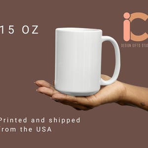 May include: A white ceramic mug with a capacity of 15 oz. The mug has a large handle and is held in a person's hand. The mug is plain and has a glossy finish. The text "DESIGN GIFTS STUDIO" is visible.