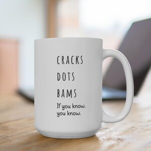 May include: White ceramic mug with the words "CRACKS DOTS BAMS" stacked vertically, followed by "If you know, you know." in black text. The mug has a large handle and is sitting on a wooden surface.