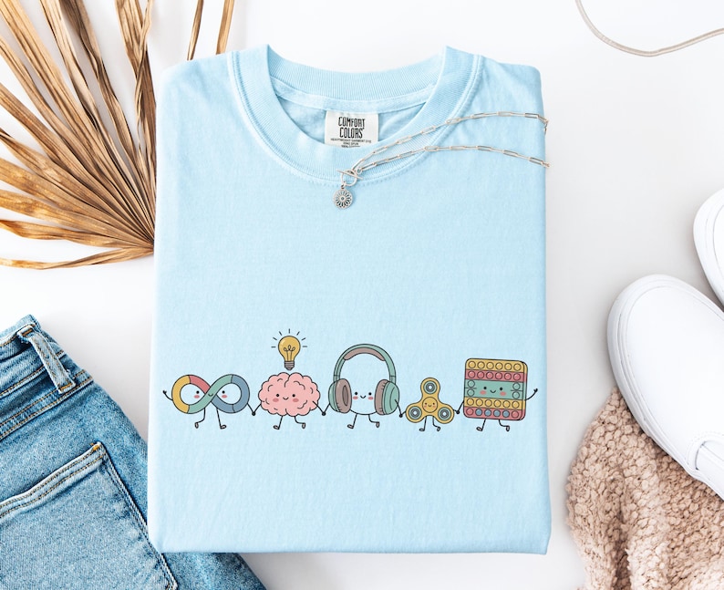 Comfort Colors Autism Doodle Characters Shirt featuring colorful, hand-drawn inclusive characters. Neatly styled next to classroom supplies, making it an ideal inclusion shirt, autism teacher shirt, and back to school shirt.