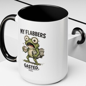 K&ouml;nnte beinhalten: Wei&szlig;er Keramikbecher mit schwarzem Innenraum, Henkel und Rand. Der Becher zeigt einen Cartoon-Frosch mit gro&szlig;en Augen, der eine Kaffeetasse und Zeitungen h&auml;lt. Der Text auf dem Becher lautet "MY FLABBERS GASTED. Daily."
