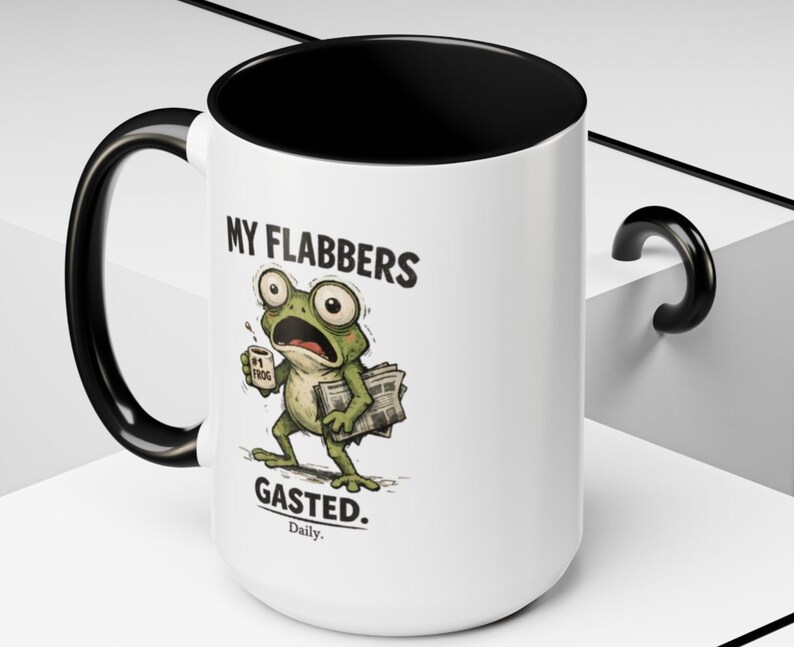 K&ouml;nnte beinhalten: Wei&szlig;er Keramikbecher mit schwarzem Innenraum, Henkel und Rand. Der Becher zeigt einen Cartoon-Frosch mit gro&szlig;en Augen, der eine Kaffeetasse und Zeitungen h&auml;lt. Der Text auf dem Becher lautet "MY FLABBERS GASTED. Daily."