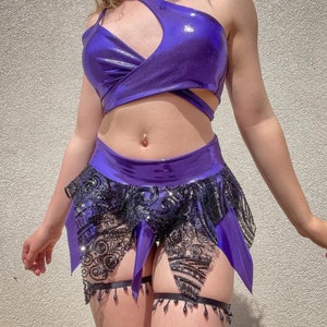Op de afbeelding: Een paars en zwart pailletten crop top en rok set. De top is een een schouder stijl met een uitsparing. De rok is een mini rok met een gerimpelde zoom.