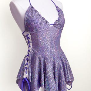 May include: A shimmering purple halter top with a flared skirt, displayed on a mannequin. The fabric has a textured, dotted pattern. The sides feature white panels with purple lacing. The top has a ruffled edge and purple ties.