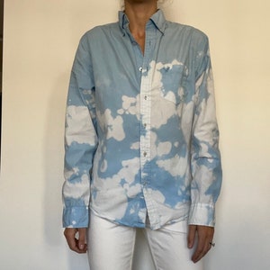 May include: A light blue button-down shirt with a white tie-dye pattern. The shirt has a single chest pocket.