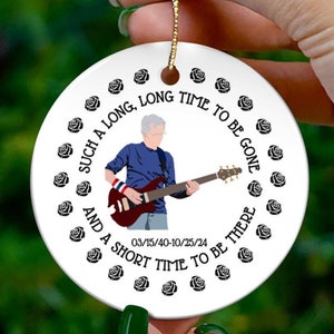May include: A white ceramic bauble featuring a black and white illustration of a person playing a guitar. The bauble is encircled by black roses and the text "SUCH A LONG, LONG TIME TO BE GONE AND A SHORT TIME TO BE THERE 03/15/40-10/25/24".