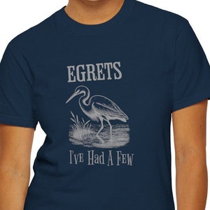 May include: Navy blue t-shirt featuring a detailed illustration of a heron in a marsh setting. The text "EGRETS I'VE HAD A FEW" is printed above and below the heron image in a light grey color. The shirt is made of a soft material.