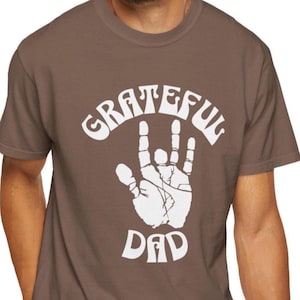 May include: A brown t-shirt with the words "GRATEFUL DAD" in white, featuring a hand gesture design. The shirt is a crew neck style and appears to be made of a soft material.