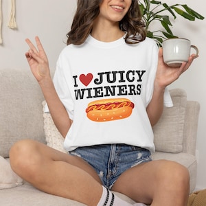 May include: A white short-sleeve t-shirt featuring the black text I, a red heart symbol, and the black text JUICY WIENERS. Below the text is a graphic illustration of a hot dog in a bun with yellow mustard and red ketchup lines on top.