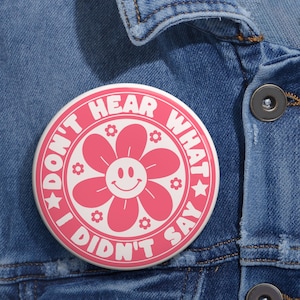 May include: A round, white button with the text "DON'T HEAR WHAT I DIDN'T SAY" in pink. The button features a pink flower with a smiling face in the center and small flower accents. The button is pinned to a blue denim jacket.