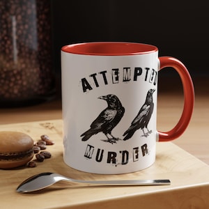 May include: White ceramic mug with a red interior and handle. The mug features the text "ATTEMPTED MURDER" above and below two black crow illustrations.
