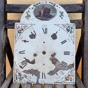 Puede incluir: Una esfera de reloj rectangular de color blanco roto con una parte superior arqueada que presenta una escena ovalada pintada de una persona sentada en una roca. La esfera muestra números romanos negros, motivos florales y desgaste.