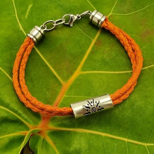 Puede incluir: Un brazalete de cuero naranja trenzado con un dije de plata con un diseño de sol.