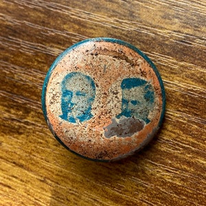 May include: A vintage metal pin with two blue and white faces on a faded orange background. The pin is slightly worn and has a blue border.