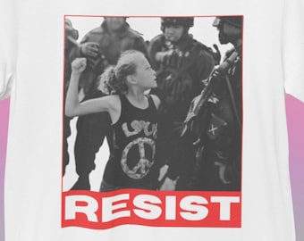 May include: White t-shirt featuring a black and white image of a young person facing a group of soldiers. The word "RESIST" is printed in red at the bottom of the image. The shirt is hanging on a wooden hanger.