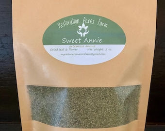 May include: A brown paper bag with a clear window, filled with dried green leaves and flowers. The bag has a green oval label with the text "Restoration Acres Farm" and "Sweet Annie". The label also includes the text "artemisia annua", "Dried leaf & flower", and "Net Weight: 2 oz".