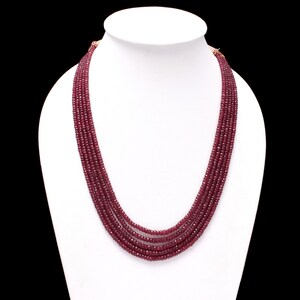 May include: A multi-strand necklace composed of small, deep red beads. The necklace is displayed on a white bust against a black background. The strands cascade down, showcasing the necklace's layered design and color.
