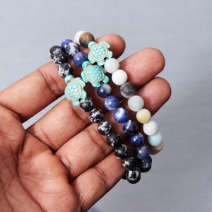 Può includere: Tre braccialetti di perline con ciondoli a forma di tartaruga turchese. I braccialetti sono sovrapposti su una mano e presentano perline nere e bianche, blu e verde chiaro.