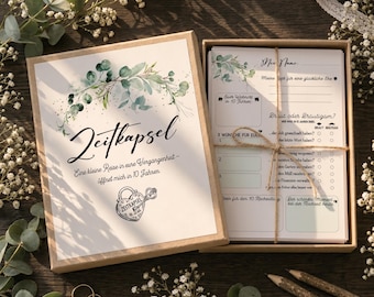 May include: A rectangular cardboard box containing a stack of printed cards tied with twine. The lid features a watercolor eucalyptus wreath, the text Zeitkapsel, and a heart-shaped padlock illustration. The cards include German text and blank lines.