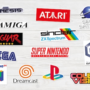 Puede incluir: Un conjunto de pegatinas coloridas con logotipos de consolas de videojuegos clásicas, incluyendo Sega Genesis, Atari, Amiga, Jaguar, Sega, Super Nintendo, Nintendo 64, Dreamcast, y otras. Las pegatinas están sobre una superficie de madera clara.