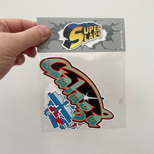 Pu&ograve; includere: Un sacchetto di plastica trasparente contenente un set di adesivi colorati con le parole "Super Slaps" e "Galaxia" stampate su di essi.