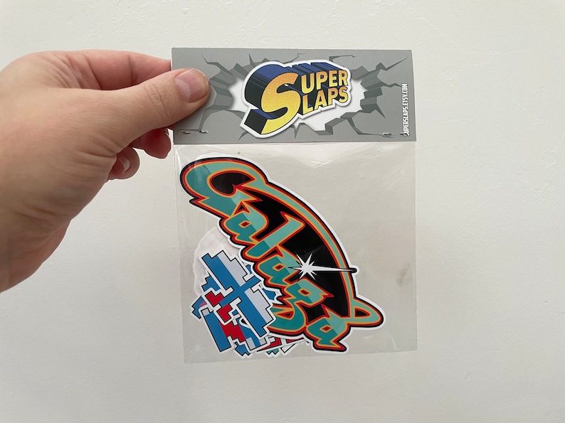 Pu&ograve; includere: Un sacchetto di plastica trasparente contenente un set di adesivi colorati con le parole "Super Slaps" e "Galaxia" stampate su di essi.