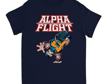 Op de afbeelding: Marineblauw T-shirt met de tekst "ALPHA FLIGHT" in rood en wit. Het shirt toont een stripboekillustratie van een personage in een handstand, met blauwe, gele en bruine tinten.